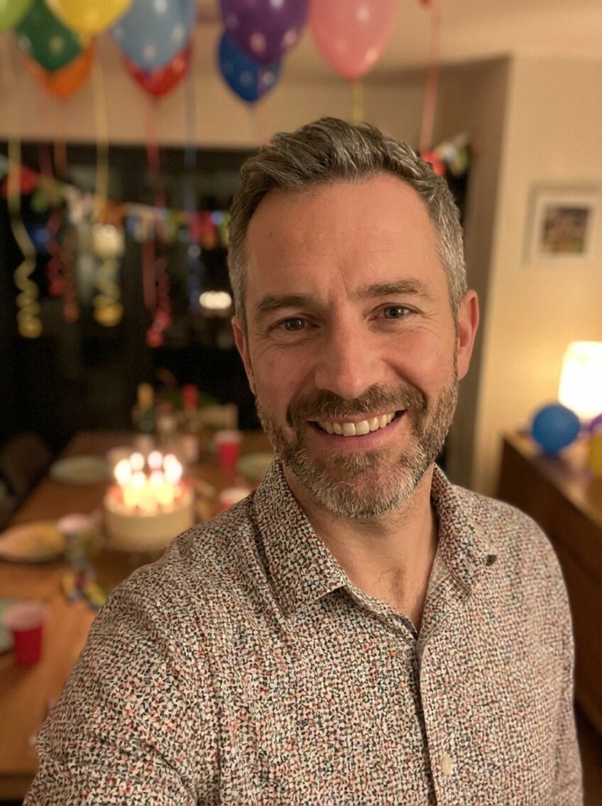A man at his home birthday dinner, a cluster of balloons and paper streamers above him and a candlelit cake glowing on the table behind