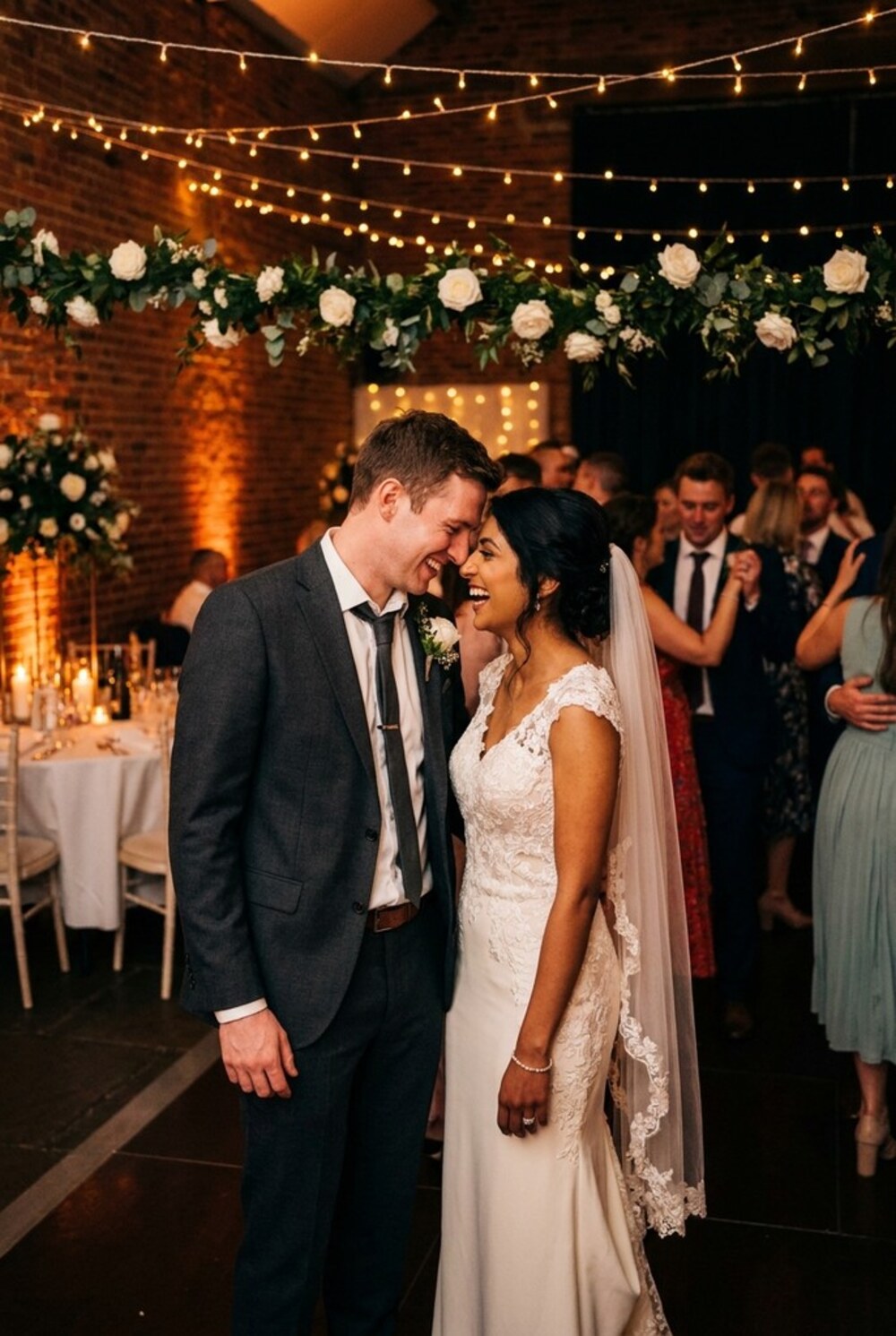A newly married couple laughing together at the edge of the dancefloor, under festoon lights and a garland of white roses